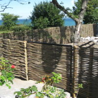 Vandret pilehegn med smedejernslåge opsat i Hellebæk. Hegnet følger terrænnet ned til vandet.